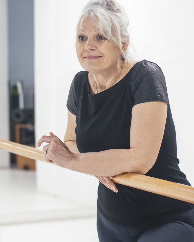 Pretty senior Caucasian woman standing by the ballet barre at gym.