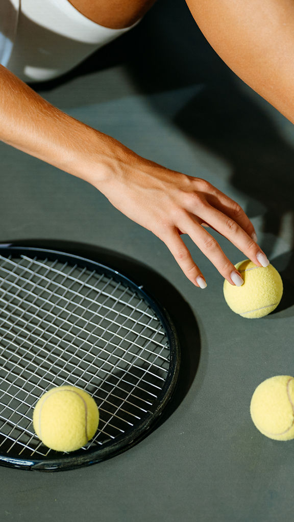 A woman in sportswear is sitting on the floor of a tennis court. A tennis racket and tennis balls are lying on the floor.