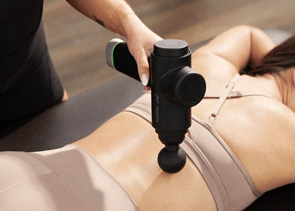 A Life Time Dynamic Stretch personal trainer using a hyperice massager on a woman's back while she lies on a table