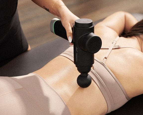 A Life Time Dynamic Stretch personal trainer using a hyperice massager on a woman's back while she lies on a table