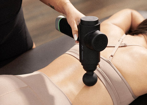 A Life Time Dynamic Stretch personal trainer using a hyperice massager on a woman's back while she lies on a table