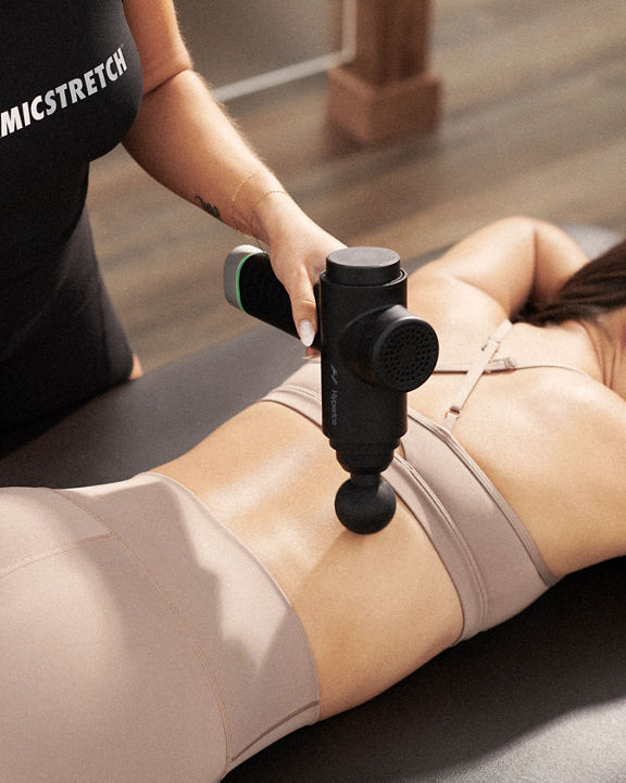 A Life Time Dynamic Stretch personal trainer using a hyperice massager on a woman's back while she lies on a table