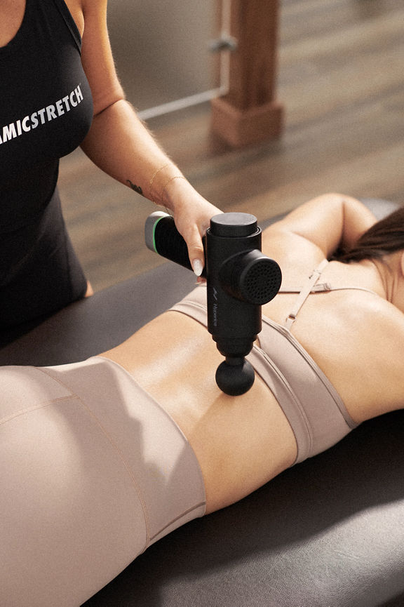 A Life Time Dynamic Stretch personal trainer using a hyperice massager on a woman's back while she lies on a table