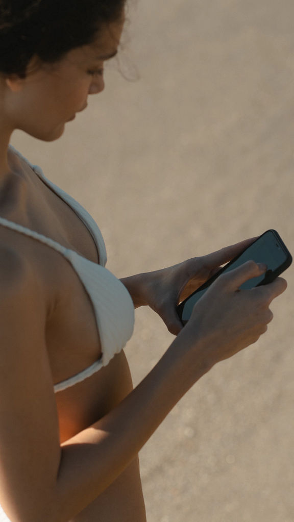An attractive female with dark braided hair standing on a sandy beach looking at her phone