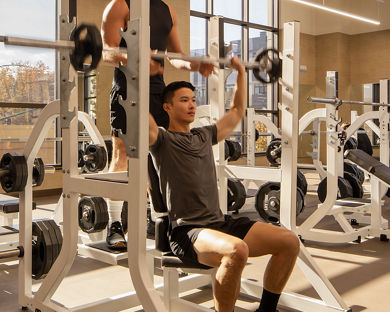 A man spotting another member while he completes overhead presses while seated on a weight bench