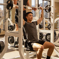 A man spotting another member while he completes overhead presses while seated on a weight bench