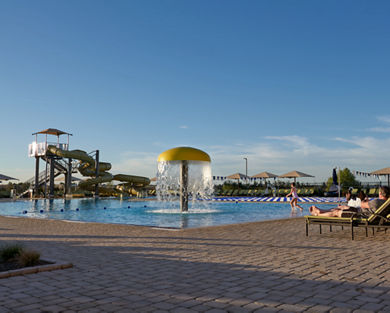 a young girl runs into an outdoor pool complete with mushroom waterfall, lap pool, waterslides, and surrounded by lounge chairs