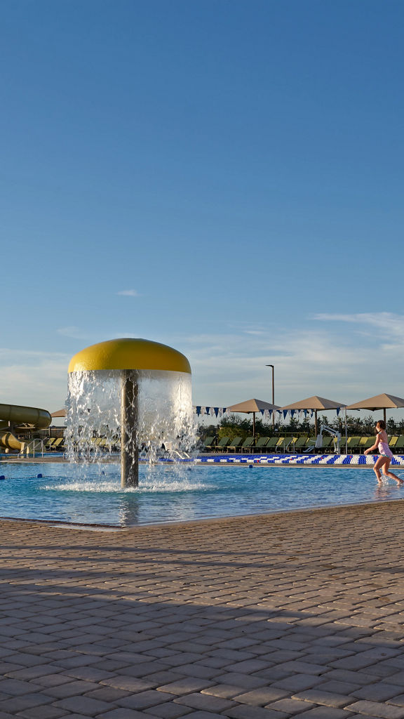 a young girl runs into an outdoor pool complete with mushroom waterfall, lap pool, waterslides, and surrounded by lounge chairs