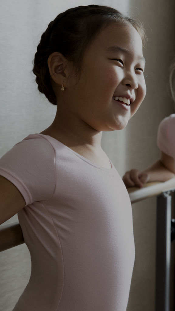 Two young girls in pink leotards smiling while standing in a studio next to a dance bar