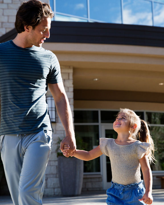 A man holds a female child’s hand as they walk together outside a Life Time building