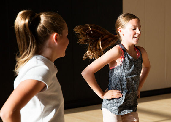 Two female children wearing athletic gear stand smiling with their hands on their hips in a Life Time gym