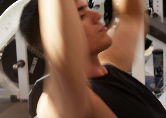 Blurred image depicting the motion of a young man lifting weights over his head while seated on a weight bench