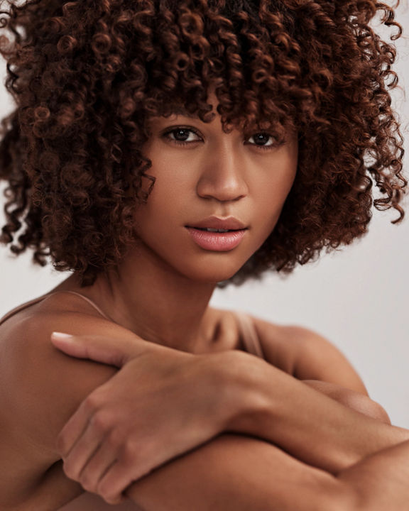 Close-up portrait of an attractive female with curly brunette hair looking directly at the camera, showcasing her flawless skin