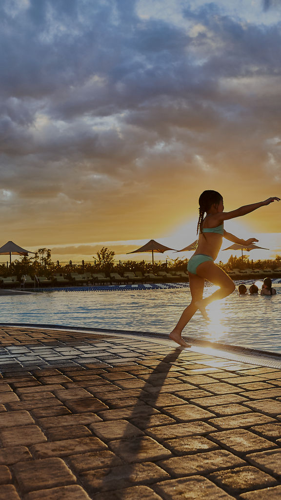 A man watches a female child jump into an outdoor pool at Life Time as the sun sets in the background