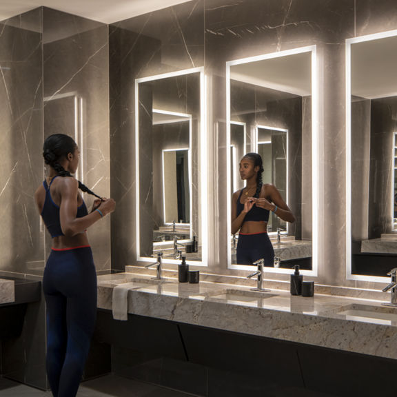 A female braiding her hair while looking into a mirror in the Life Time One Wall locker room