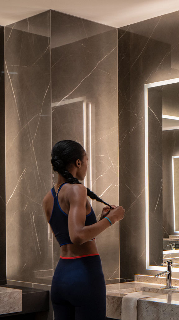 A female braiding her hair while looking into a mirror in the Life Time One Wall locker room