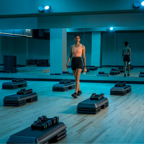 A woman walks through rows of equipment in a blue lit group fitness studio