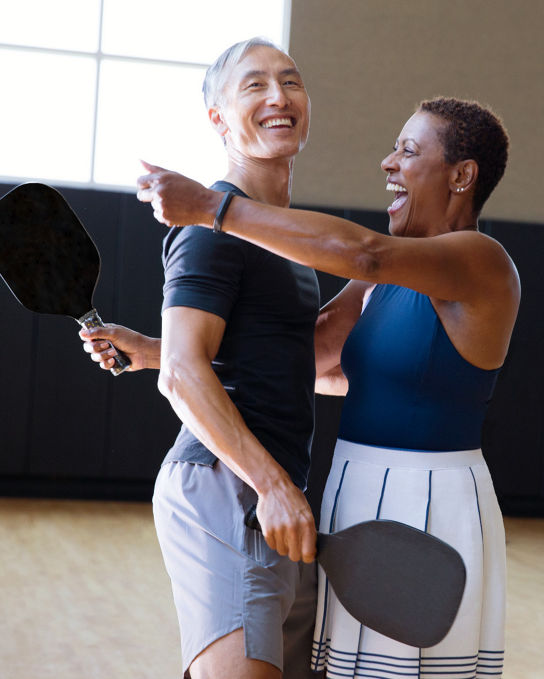 A man and a woman each holding a pickleball racuqet about to embrace eachother while standing on an indoor pickleball court