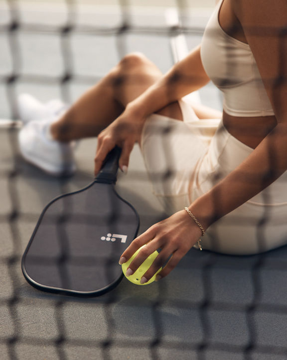 An adult female holding a pickleball paddle and ball while sitting next to a net