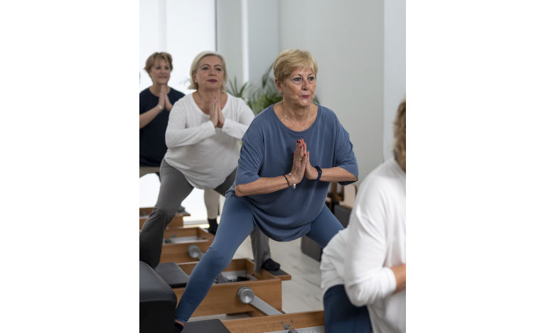 Group of concentrated mature women in sportswear performing side splits exercise on reformer machine while practicing Pilates in modern light studio.