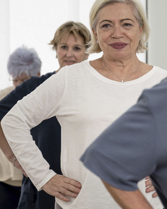 Group of happy senior ladies in activewear standing on reformers with hands on waists during Pilates training in modern light studio.