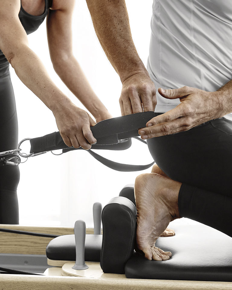 A man using a Pilates reformer machine with a Life Time trainer