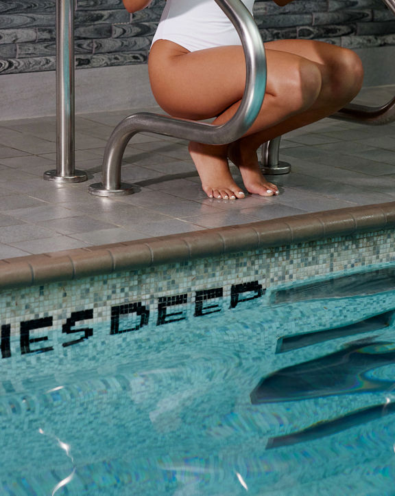 Woman wearing a white bathing suit kneeling near an indoor pools edge; looking off-camera