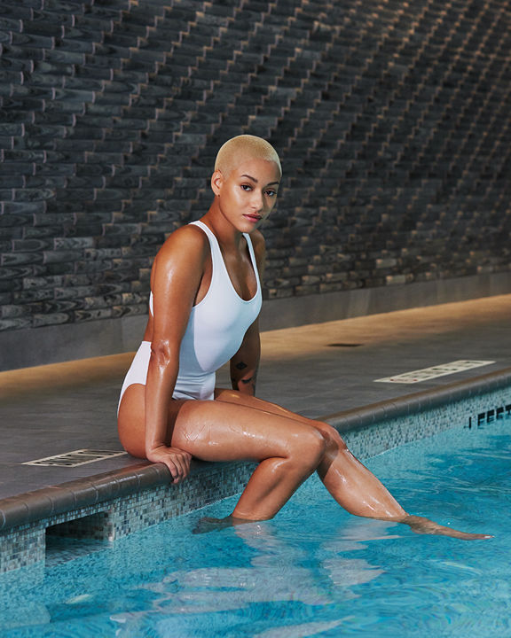 Woman wearing a white bathing suit sitting on an indoor pools edge dipping her feet into the water; looking directly at the camera