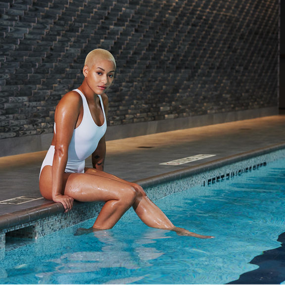 Woman wearing a white bathing suit sitting on an indoor pools edge dipping her feet into the water; looking directly at the camera