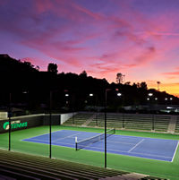 An outdoor tennis court lit by lights in the evening, dramatic colorful sky in the background