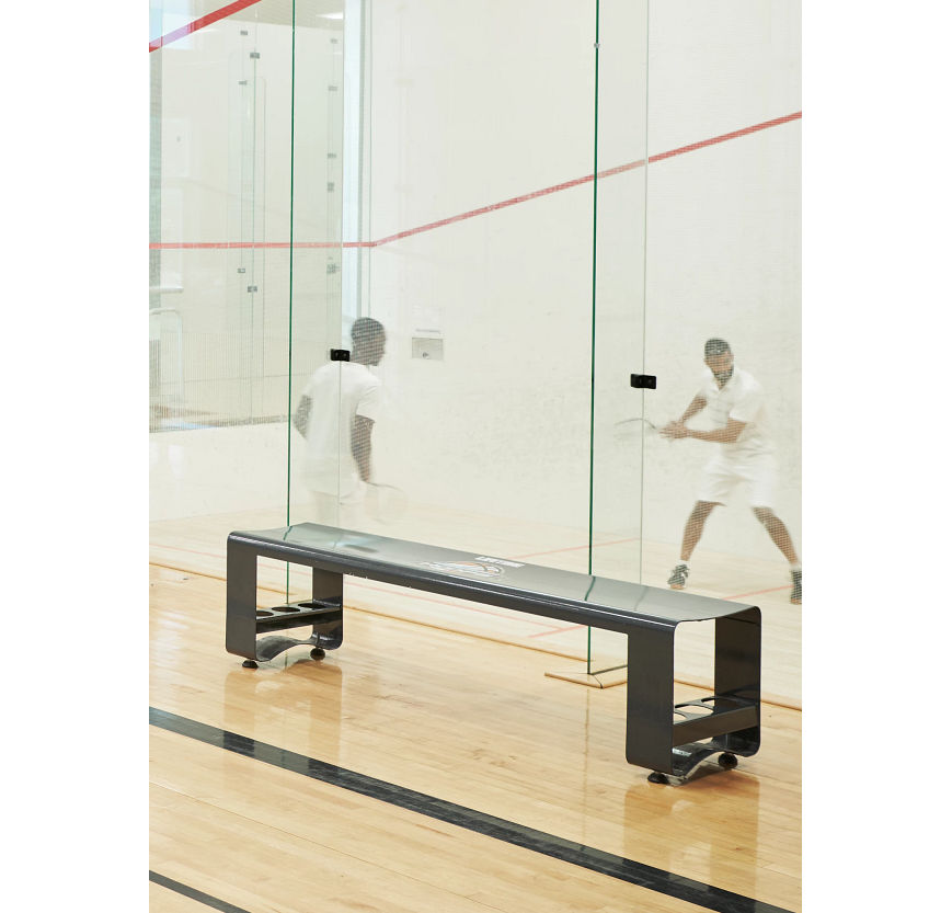 A woman walking by a squash court while two men are playing a squash match