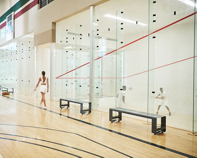 A woman walking by a squash court while two men are playing a squash match