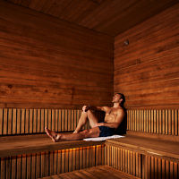 Man sitting in a sauna with his legs out and leaning against the wall with his eyes closed.