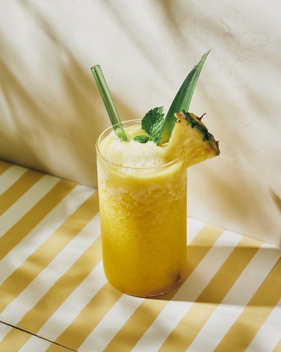 A pineapple smoothie garnished with a fresh slice of pineapple and mint leaf placed on a yellow and white striped tabletop