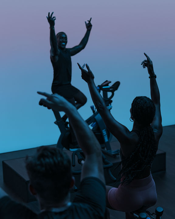 AMP spin class riding together, led by their instructor, with their hands in the air