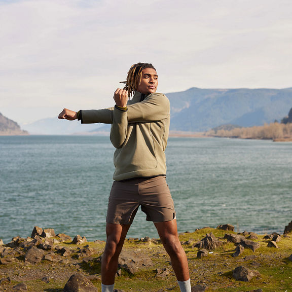 An adult male, standing next to water with mountains in the background, stretching his arms