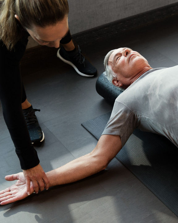 A man lying on his back on a yoga mat while a trainer helps him stretch