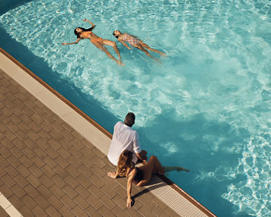 Mother and father sitting on the edge of an outdoor pool watching their children swim