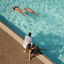 Mother and father sitting on the edge of an outdoor pool watching their children swim