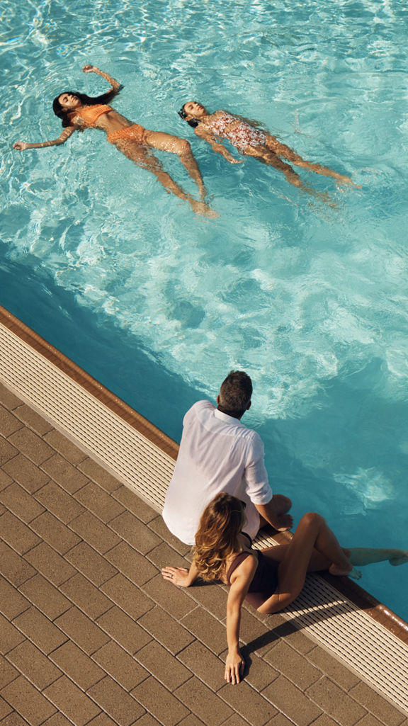 Mother and father sitting on the edge of an outdoor pool watching their children swim