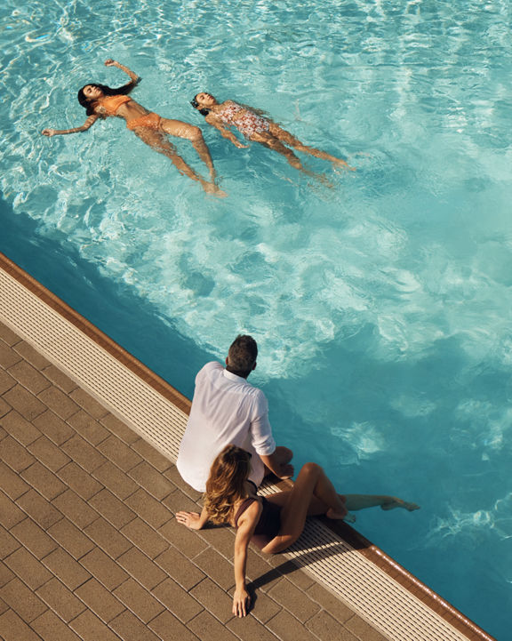 Mother and father sitting on the edge of an outdoor pool watching their children swim