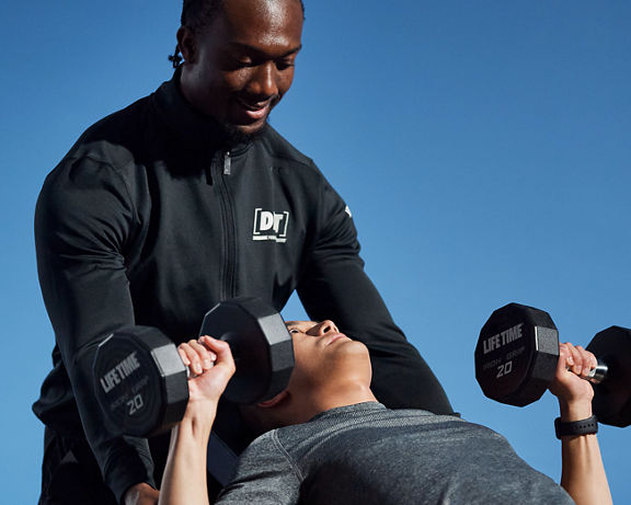 A male Life Time Dynamic Personal Trainer providing assistance while a participant completes a chest press using dumbbells