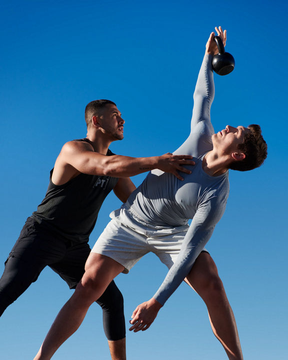 A Life Time Dynamic Personal Trainer helping adjust body positioning as a male member holds a kettlebell above his head