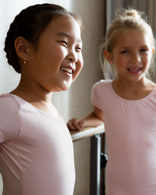 Two young girls in pink leotards smiling while standing in a studio next to a dance bar
