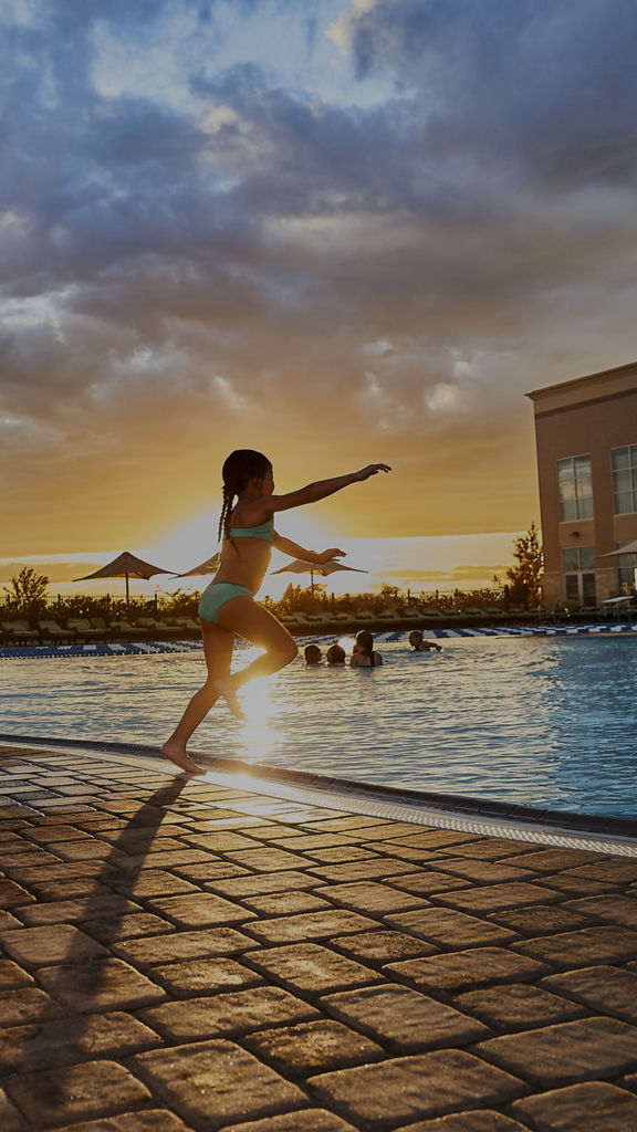A man watches a female child jump into an outdoor pool at Life Time as the sun sets in the background