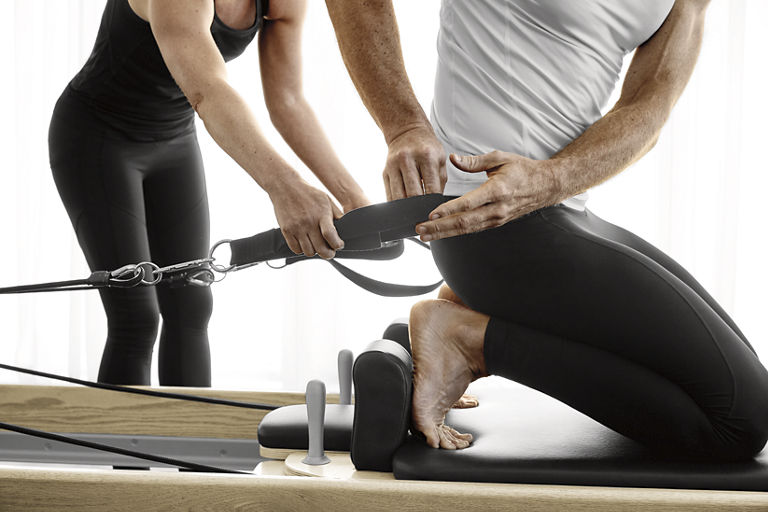 A man using a Pilates reformer machine with a Life Time trainer