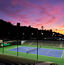 An outdoor tennis court with stadium seating lit by lights in the evening, dramatic colorful sky in the background
