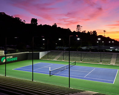 An outdoor tennis court with stadium seating lit by lights in the evening, dramatic colorful sky in the background