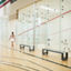 A woman walking by a squash court while two men are playing a squash match